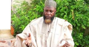 Unpaid N9m Debt: Alhaji Akewugbagold To Face Legal Action Man in beige traditional robe and patterned cap gestures with his hands outdoors, green bushes in the background.