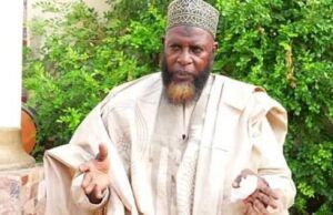 Unpaid N9m Debt: Alhaji Akewugbagold To Face Legal Action Man in beige traditional robe and patterned cap gestures with his hands outdoors, green bushes in the background.