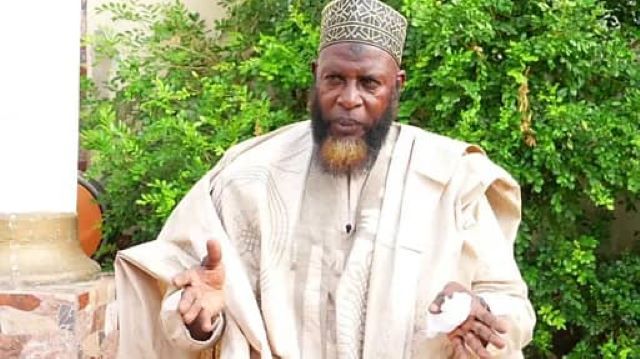 Akewugagold Man in beige traditional robe and patterned cap gestures with his hands outdoors, green bushes in the background.
