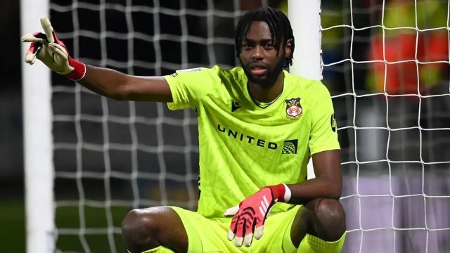 Arthur Okonkwo Soccer goalkeeper in a neon yellow kit crouches in front of the goal, wearing red gloves and pointing with one arm at the left side.