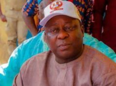 Osun Accord Chairman Shot, Police Begin Probe Man wearing a beige traditional outfit and a white cap sits with a blue water bottle nearby, looking to the side.