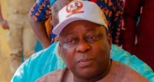 Osun Accord Chairman Shot, Police Begin Probe Man wearing a beige traditional outfit and a white cap sits with a blue water bottle nearby, looking to the side.