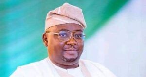 Barr Akeem Agbaje Condemns Abduction Of Adelabu’s Sister, Twin Sons Smiling man in white traditional attire and cap with glasses, posed against a green backdrop.