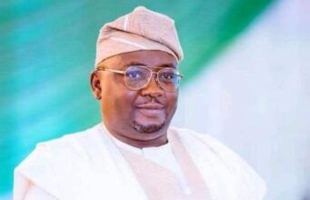 Barr Akeem Agbaje Condemns Abduction Of Adelabu’s Sister, Twin Sons Smiling man in white traditional attire and cap with glasses, posed against a green backdrop.