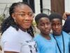 Alli Condemns Ex-Minister Adelabu’s Sister’s Abduction, Urges Swift Rescue Three people posing for a photo outside a building: an adult woman with glasses and two young boys in blue shirts, all smiling at the camera.