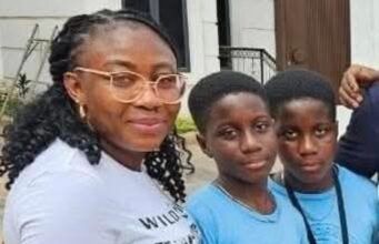 Alli Condemns Ex-Minister Adelabu’s Sister’s Abduction, Urges Swift Rescue Three people posing for a photo outside a building: an adult woman with glasses and two young boys in blue shirts, all smiling at the camera.