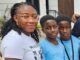 Alli Condemns Ex-Minister Adelabu’s Sister’s Abduction, Urges Swift Rescue Three people posing for a photo outside a building: an adult woman with glasses and two young boys in blue shirts, all smiling at the camera.