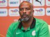 Playing Against Super Eagles Is A Big Opportunity For Portugal – Chelle Man wearing a green Nigeria football polo at a press conference, speaking into a microphone with a backdrop of logos behind him.