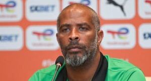 Chelle Gets New Improved Eagles Contract, Handed Olympic Eagles Job Man wearing a green Nigeria football polo at a press conference, speaking into a microphone with a backdrop of logos behind him.