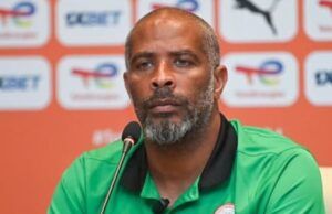 Playing Against Super Eagles Is A Big Opportunity For Portugal – Chelle Man wearing a green Nigeria football polo at a press conference, speaking into a microphone with a backdrop of logos behind him.