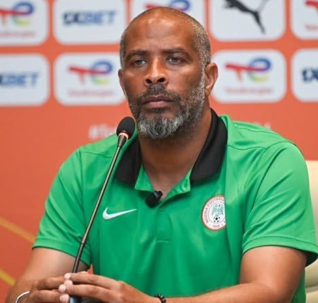 Eric Chelle Man wearing a green Nigeria football polo at a press conference, speaking into a microphone with a backdrop of logos behind him.