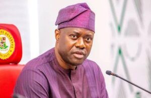 Pity Fayose, Pray For Him, Makinde Tells Nigerians Man in a purple traditional outfit and cap speaks into a microphone at a formal meeting, seated behind a desk with pens in his chest pocket.