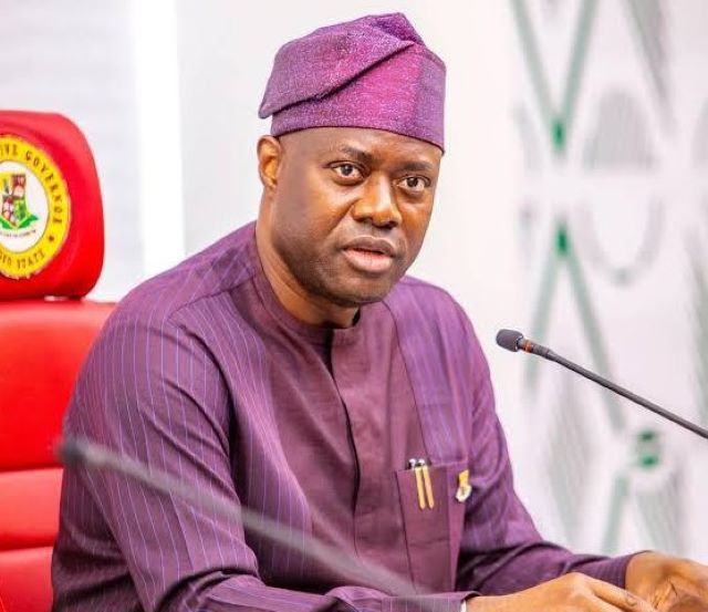 Gov Makinde new Man in a purple traditional outfit and cap speaks into a microphone at a formal meeting, seated behind a desk with pens in his chest pocket.