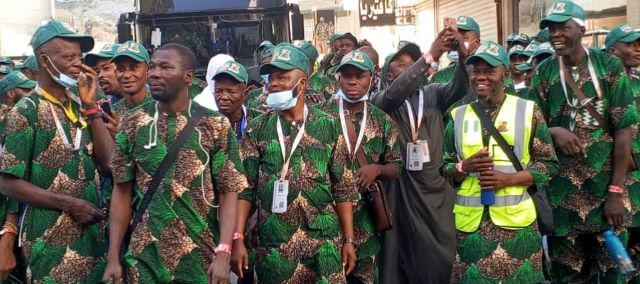 Hajj Return Group of men in matching green camouflage outfits and green caps posing together, some with lanyards and a high-visibility vest in the crowd.