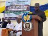 NOA DG Charges Nigerians To Combine Artificial Intelligence With Values, Productivity For National Development Speaker at a wooden podium with a blue-and-gold bow delivering a talk at a conference; a banner behind shows event details and logos.