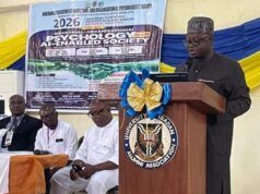 NOA DG Charges Nigerians To Combine Artificial Intelligence With Values, Productivity For National Development Speaker at a wooden podium with a blue-and-gold bow delivering a talk at a conference; a banner behind shows event details and logos.