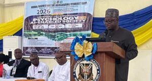 NOA DG Charges Nigerians To Combine Artificial Intelligence With Values, Productivity For National Development Speaker at a wooden podium with a blue-and-gold bow delivering a talk at a conference; a banner behind shows event details and logos.