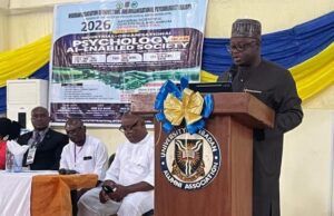 NOA DG Charges Nigerians To Combine Artificial Intelligence With Values, Productivity For National Development Speaker at a wooden podium with a blue-and-gold bow delivering a talk at a conference; a banner behind shows event details and logos.