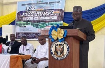 NOA DG Charges Nigerians To Combine Artificial Intelligence With Values, Productivity For National Development Speaker at a wooden podium with a blue-and-gold bow delivering a talk at a conference; a banner behind shows event details and logos.