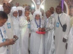 Foundation Installs Yeye Omikunle As Patron, Offers Prayers For Abducted Oriire Students, Teachers Group of people in white religious robes at an indoor ceremony; a woman holding a microphone stands center, clasping hands with an older man while others look on.