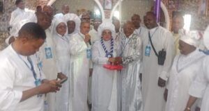 Foundation Installs Yeye Omikunle As Patron, Offers Prayers For Abducted Oriire Students, Teachers Group of people in white religious robes at an indoor ceremony; a woman holding a microphone stands center, clasping hands with an older man while others look on.
