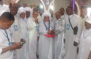 Foundation Installs Yeye Omikunle As Patron, Offers Prayers For Abducted Oriire Students, Teachers Group of people in white religious robes at an indoor ceremony; a woman holding a microphone stands center, clasping hands with an older man while others look on.