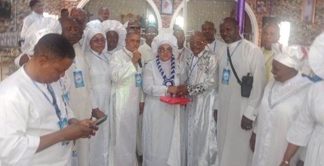 Mrs Omikunle Group of people in white religious robes at an indoor ceremony; a woman holding a microphone stands center, clasping hands with an older man while others look on.