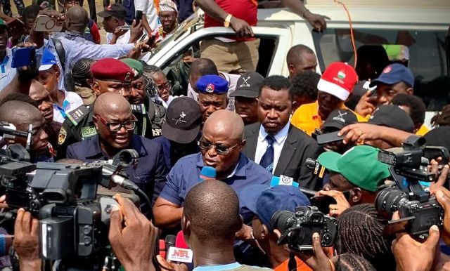 Obasa Politician or speaker addressing a crowded media scrum with multiple cameras and microphones around him near a white vehicle.