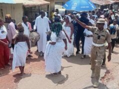 Oyo School Abduction: Ogun Traditionalists Stage Procession, Demand Safe Release Of Kidnapped Student, Teachers Dancers in white walk and circle on a dusty street as a drummer plays; a uniformed officer points directions to the crowd nearby under sunny skies.