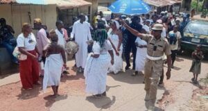 Oyo School Abduction: Ogun Traditionalists Stage Procession, Demand Safe Release Of Kidnapped Student, Teachers Dancers in white walk and circle on a dusty street as a drummer plays; a uniformed officer points directions to the crowd nearby under sunny skies.