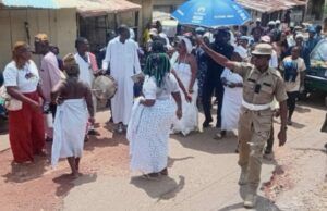 Oyo School Abduction: Ogun Traditionalists Stage Procession, Demand Safe Release Of Kidnapped Student, Teachers Dancers in white walk and circle on a dusty street as a drummer plays; a uniformed officer points directions to the crowd nearby under sunny skies.