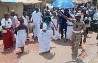 Oyo School Abduction: Ogun Traditionalists Stage Procession, Demand Safe Release Of Kidnapped Student, Teachers Dancers in white walk and circle on a dusty street as a drummer plays; a uniformed officer points directions to the crowd nearby under sunny skies.