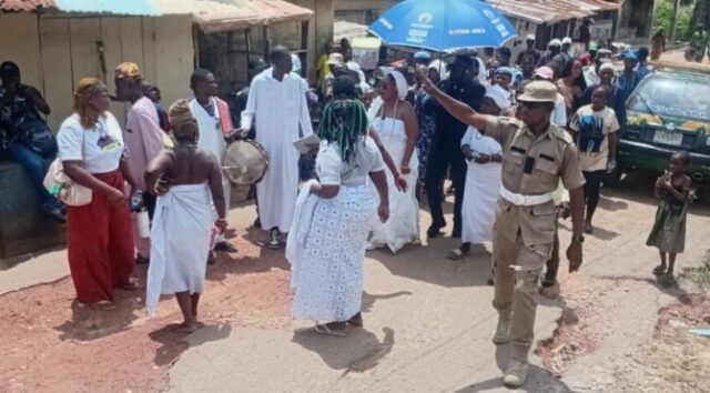 Dancers in white walk and circle on a dusty street as a drummer plays; a uniformed officer points directions to the crowd nearby under sunny skies.