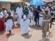 Oyo School Abduction: Ogun Traditionalists Stage Procession, Demand Safe Release Of Kidnapped Student, Teachers Dancers in white walk and circle on a dusty street as a drummer plays; a uniformed officer points directions to the crowd nearby under sunny skies.
