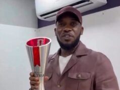 After 28 Years: Okocha Gets Retro 1998 World Cup Player Of Match Award Man in a brown jacket and cap holding a tall silver trophy with a red ribbon indoors, smiling for the camera.