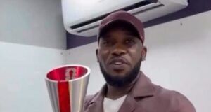 After 28 Years: Okocha Gets Retro 1998 World Cup Player Of Match Award Man in a brown jacket and cap holding a tall silver trophy with a red ribbon indoors, smiling for the camera.