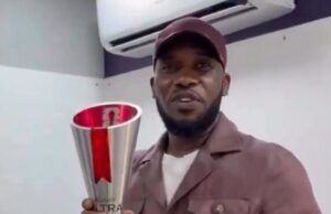 After 28 Years: Okocha Gets Retro 1998 World Cup Player Of Match Award Man in a brown jacket and cap holding a tall silver trophy with a red ribbon indoors, smiling for the camera.