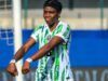 Oshoala Sends Message To Tinubu After Nigeria’s Win Over Senegal Female soccer player in a green-and-white striped kit celebrating with arms outstretched on the field, goalpost and blue barriers in the background.