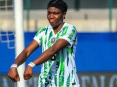 Oshoala Sends Message To Tinubu After Nigeria’s Win Over Senegal Female soccer player in a green-and-white striped kit celebrating with arms outstretched on the field, goalpost and blue barriers in the background.
