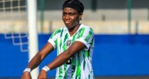 Oshoala Sends Message To Tinubu After Nigeria’s Win Over Senegal Female soccer player in a green-and-white striped kit celebrating with arms outstretched on the field, goalpost and blue barriers in the background.