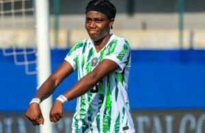 Oshoala Sends Message To Tinubu After Nigeria’s Win Over Senegal Female soccer player in a green-and-white striped kit celebrating with arms outstretched on the field, goalpost and blue barriers in the background.