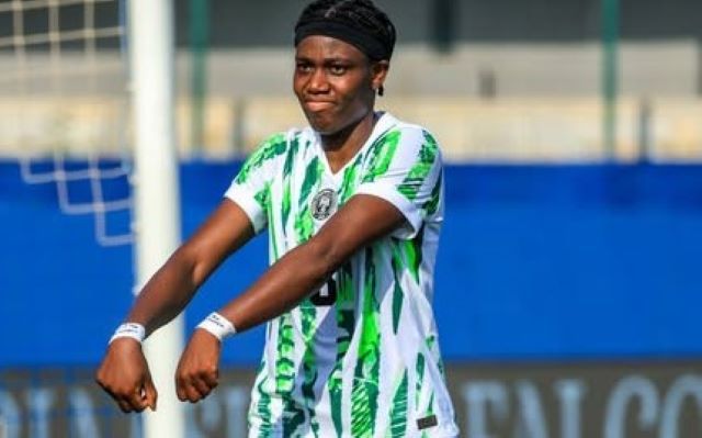 Oshoala message Female soccer player in a green-and-white striped kit celebrating with arms outstretched on the field, goalpost and blue barriers in the background.