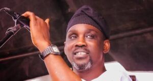Pasuma Threatens Lawsuit Over Unauthorised Copyright Infringement Man on stage speaking into a microphone, wearing a black head wrap and white shirt, smiling at the audience.