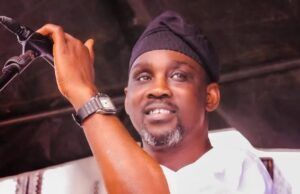 Alabi Pasuma Threatens Lawsuit Over Copyright Infringement Man on stage speaking into a microphone, wearing a black head wrap and white shirt, smiling at the audience.