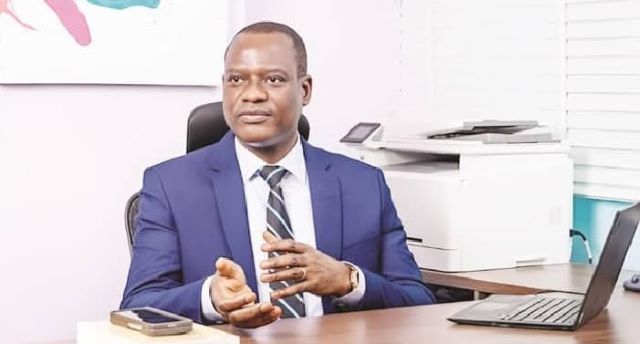 Taiwo Oyedele Professional man in a blue suit seated at a desk in an office, gesturing with his hands as he speaks.