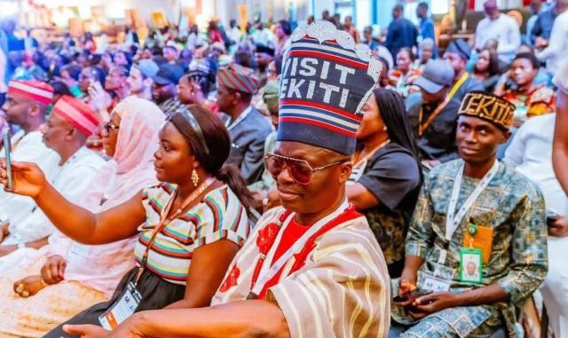 Visit-Ekiti-Bill-board-cap-Gains-Shettima Crowd of people at a formal gathering, many wearing traditional hats and lanyards, seated in rows.